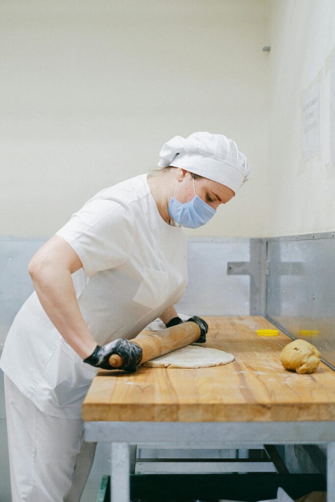 A masked pastry chef uses a rolling pin to flatten dough on a wooden surface in a kitchen.
